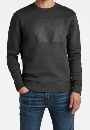 Mand iført mørkegrå sweatshirt med præget "RAW" tekst og blå jeans, hænderne i lommerne, stående mod en ensfarvet baggrund.