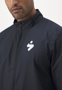 Marine vindjakke med høy krage, glidelås foran og hvit logo på venstre bryst; glatt tekstur, lett materiale.