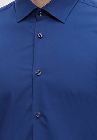 Gros plan d'une personne portant une chemise habillée bleu foncé boutonnée avec un col pointu.