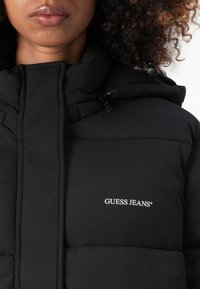 Black puffer jacket with a high collar, featuring a branded logo in white on the chest and textured quilted design.