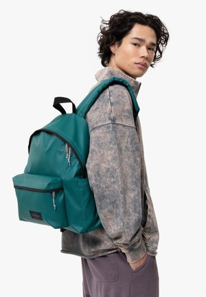 Sac à dos teal fabriqué à partir d'un matériau durable, avec une poche frontale, des accents de zip noir et des bretelles rembourrées, présenté sur un modèle.