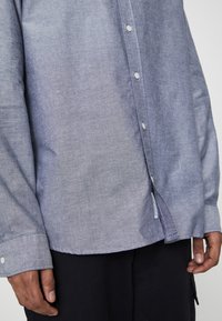 Gros plan sur une personne portant une chemise bleue à boutons blancs et un pantalon noir, les mains détendues le long du corps.