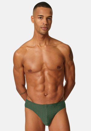 Muskelbepackter Mann mit kurzen Haaren, der grün-weiß vertikal gestreifte Slips trägt und mit den Händen hinter dem Rücken vor einem einfarbigen Hintergrund steht.