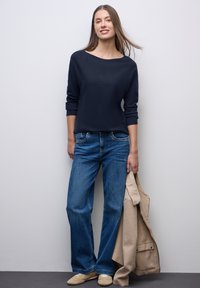 Marineblauer Pullover mit weitem Rundhalsausschnitt, blaue Jeans und beiger Mantel in einer Hand gehalten, ergänzt mit hellbeigen Schuhen. Gerade Passform und lockerer Schnitt.