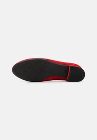 Chaussure en daim rouge avec un dessus lisse et une semelle en caoutchouc noir présentant un motif tourbillonnant. Le talon est légèrement rembourré et a une forme arrondie.