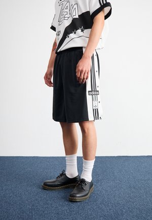 Personne portant un short Adidas noir et blanc avec des logos, des chaussures noires, des chaussettes blanches et un t-shirt blanc à motif, debout sur un tapis bleu.