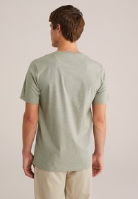 Tânăr cu păr scurt castaniu, îmbrăcat cu un tricou deschis la culoare, cu mânecă scurtă, și pantaloni bej, stând cu fața îndreptată spre o fundal simplu.