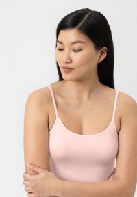 Jonge vrouw met lang donker haar, gekleed in een lichtroze topje, kijkt naar links beneden, met haar armen zachtjes over haar lichaam gekruist.