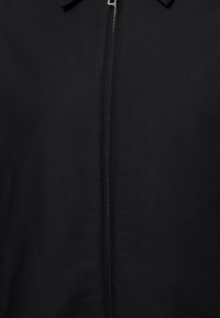 Veste noire avec une fermeture éclair verticale argentée centrée, partiellement fermée, et un petit col visible en haut.