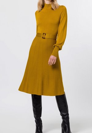 Robe tricotée jaune moutarde avec des manches longues et bouffantes, un corsage ajusté et une jupe évasée accentuée par une large ceinture et une boucle carrée.