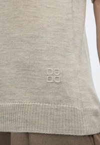 Μπεζ πλεκτό στρίφωμα πουλόβερ με μικρό κεντητό μοτίβο τετράγωνου πάνω από την ριμπάτη άκρη, φορεμένο πάνω από καφέ παντελόνι.