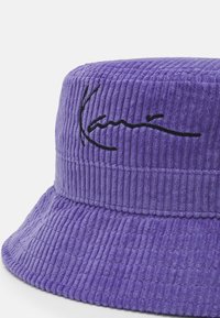 Pălărie de tip bucket din corduroy violet, cu o bordură largă și un semn distinctiv negru brodat. Materialul are dungi verticale pentru textură.