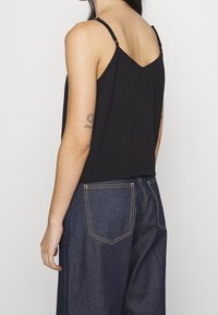 Černý tílko bez rukávů s výstřihem do V, s tenkými nastavitelnými ramínky, v kombinaci s tmavými džínami se zřetelným prošíváním a kapsami.