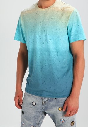 Homme portant un t-shirt à manches courtes dégradé du beige au bleu avec de petites taches et un jean clair avec des patchs circulaires colorés.