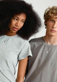 Due modelle indossano t-shirt a manica corta: una in grigio chiaro, l'altra in grigio. Entrambe indossano collane d'argento, che mostrano un design minimalista.