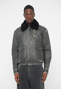 Veste en denim noir avec un col en fausse fourrure, deux poches avant, et un détail zippé, présentant une finition usée et des coutures blanches contrastantes.