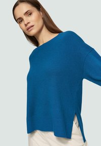 Blauwe gebreide trui met een losse pasvorm, zijsplitten en een brede halslijn. Zachte textuur met een geribde zoomafwerking.