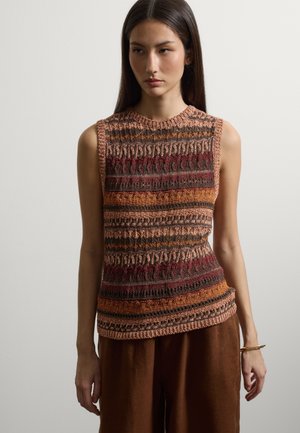 Frau trägt ärmelloses Strickoberteil mit horizontalen Streifenmustern in Braun-, Rost-, Rot- und Orangetönen, kombiniert mit locker sitzenden braunen Hosen.