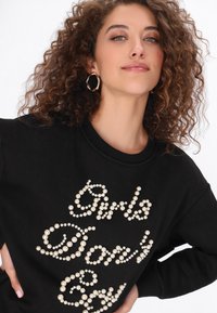 Fekete pulóver gyöngy díszítéssel, amely a "Girls Don't" feliratot formázza az elején. Kerek nyakkivágással és hosszú ujjal rendelkezik.