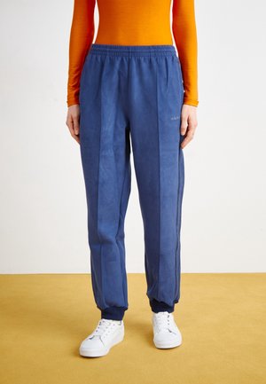 Osoba ubrana w niebieskie spodnie dresowe Adidas z elastycznymi mankietami przy kostkach oraz białe trampki, stojąca na żółtej podłodze na tle białej ściany.