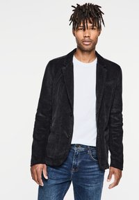 Svart kordfläta blazer med slag, två knappar och framfickor, i kombination med en vit t-shirt och blå denimjeans.