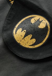 Schwarzer Baumwollstoff mit einem verwitterten gelben Batman-Logo, kreisförmige Form mit Fledermausflügel-Akzenten, auf einer leicht strukturierten Oberfläche.