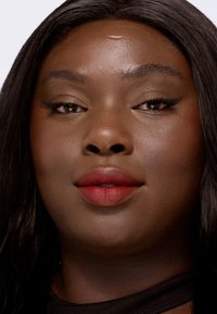 Nært portrett av et ansikt med glatt, mørk hud, fyldige lepper i rødt, mandelformede øyne og langt svart hår. Minimalistisk bakgrunn fremhever trekkene.