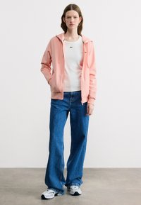 Νεαρή γυναίκα όρθια, φορώντας ροζ φούτερ με φερμουάρ, λευκό πουκάμισο, φαρδιά μπλε τζιν και λευκά αθλητικά παπούτσια, σε απλό λευκό φόντο.