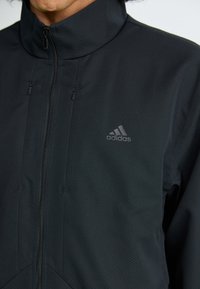 Sort atletisk jakke lavet af tekstureret stof med høj krave, lynlås fortil og to lommer med lynlås; har et diskret Adidas-logo.