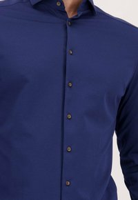 Close-up van een persoon in een getailleerd blauw overhemd met bruine knopen en een standaard kraag.