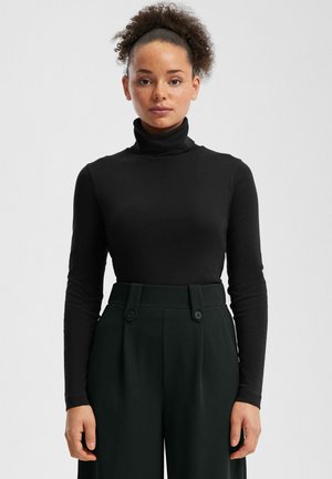 Woman with curly hair in a high puff wears a black turtleneck and high-waisted black pants, standing against a plain light background.