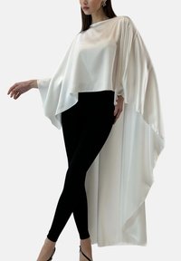Haut cape en soie blanche à design drapé, avec col montant et ourlet asymétrique, porté sur un legging noir et assorti à des talons à brides.
