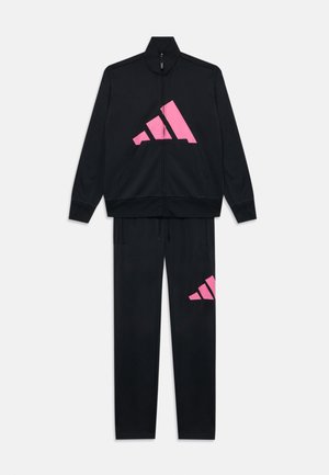 Črn trenirko, ki obsega zadrgo jakno in usklajene hlače, ki imata velike roza akcentne logotipe Adidas na obeh delih.
