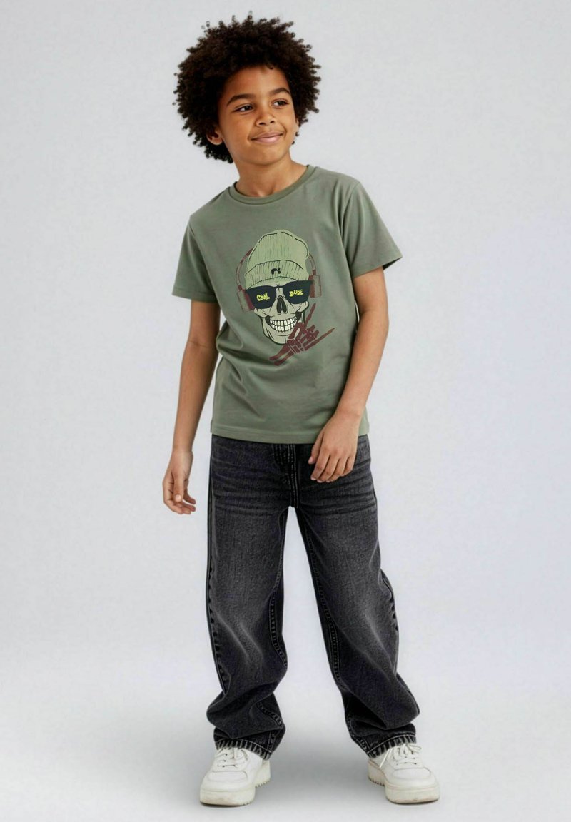 Grön bomullst-shirt med en skalle med hörlurar som motiv. Kombinerad med lediga mörkgrå jeans och vita sneakers.