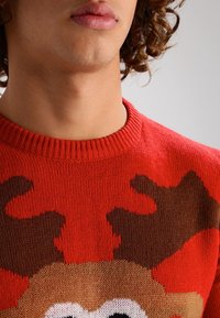 Pull rouge en tricot avec un motif de renne, présentant des bois bruns et des détails du visage. Col côtelé et texture douce.