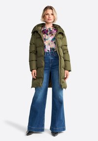 Olijfgroene gewatteerde jas met capuchon, met een ritsluiting en grote zakken, gedragen over een bloes met bloemenprint en wijde blauwe jeans.
