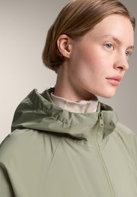 Jachetă cu glugă de culoare verde deschis, cu o textură netedă și deschidere cu fermoar pe față. Prezintă un guler înalt și elemente de design minimaliste.
