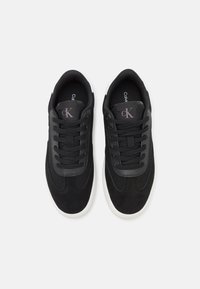 Fekete sportcipők velúr felsőrésszel és textil díszítésekkel, fehér gumi talppal és fekete fűzővel. Logó a nyelvén.