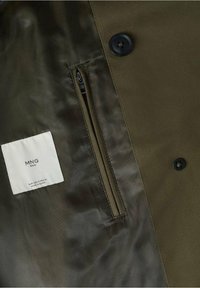 Intérieur d'une veste vert olive montrant une poche zippée, deux boutons noirs, et une étiquette blanche de la marque MNG Man avec les détails de taille et d'origine.