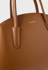 Borsa a mano in pelle marrone con manici superiori arrotondati e logo oro "COCCINELLE" impresso vicino al bordo.