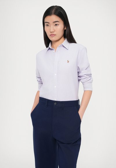 Jasnofioletowa koszula zapinana na guziki z podwiniętymi rękawami, ozdobiona małym, haftowanym logo. Do tego granatowe spodnie z bocznymi kieszeniami.