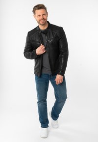 Giubbotto di pelle nera con tasche sul petto, spalle testurizzate, t-shirt grigia, jeans blu e sneakers bianche. Modello in piedi su uno sfondo neutro.
