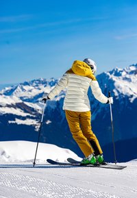 Skikleding bestaat uit een witte gewatteerde jas met een gele capuchon, gele broek, groene skibotten en skistokken, tegen de achtergrond van een besneeuwde berg.