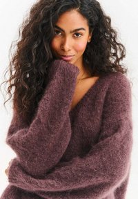 Fioletowy, puszysty sweter dzianinowy z dekoltem w serek i oversize'owymi rękawami. Miękka tekstura o luźnym kroju, podkreślająca linię szyi.
