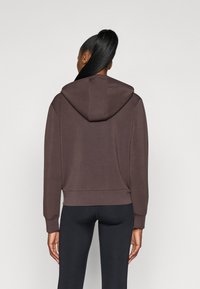 Καφέ κοντό φούτερ με κουκούλα και κορδόνι, λεία υφή, μακριά μανίκια και ribbed μανσέτες, συνδυασμένο με μαύρα κολάν, προβαλλόμενο από πίσω.