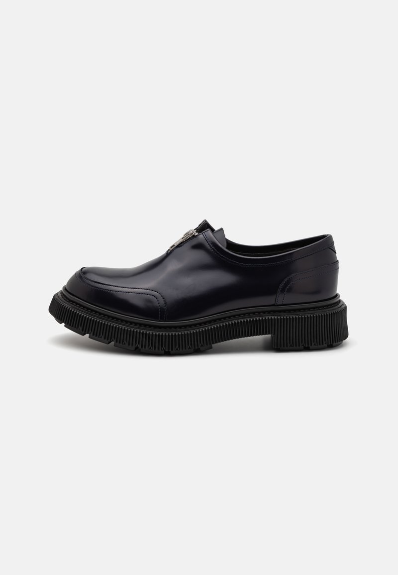 Black leather slip-on shoe with a rounded toe, textured rubber sole, and silver zipper detail on the upper. Smooth finish and minimal seams.