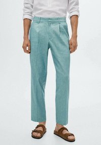 Türkisfarbene Leinenhose mit weitem Schnitt, flacher Vorderseite und zwei Fronttaschen. Kombiniert mit braunen Sandalen, die ein entspanntes Design zeigen.