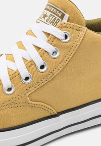 Žluté plátěné tenisky s bílými tkaničkami, kovovými průchody a černou gumovou podrážkou. Obsahují vyztuženou špičku a logo "Converse All Star".