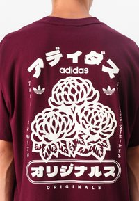 Maroon cotton t-shirt featuring a white floral graphic print, large brand logo, and text in Japanese and English on the back.