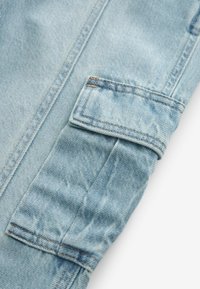 Dettaglio di una giacca di jeans azzurro chiaro con un polsino piegato e cuciture visibili. Il tessuto ha un aspetto leggermente testurizzato e usurato.